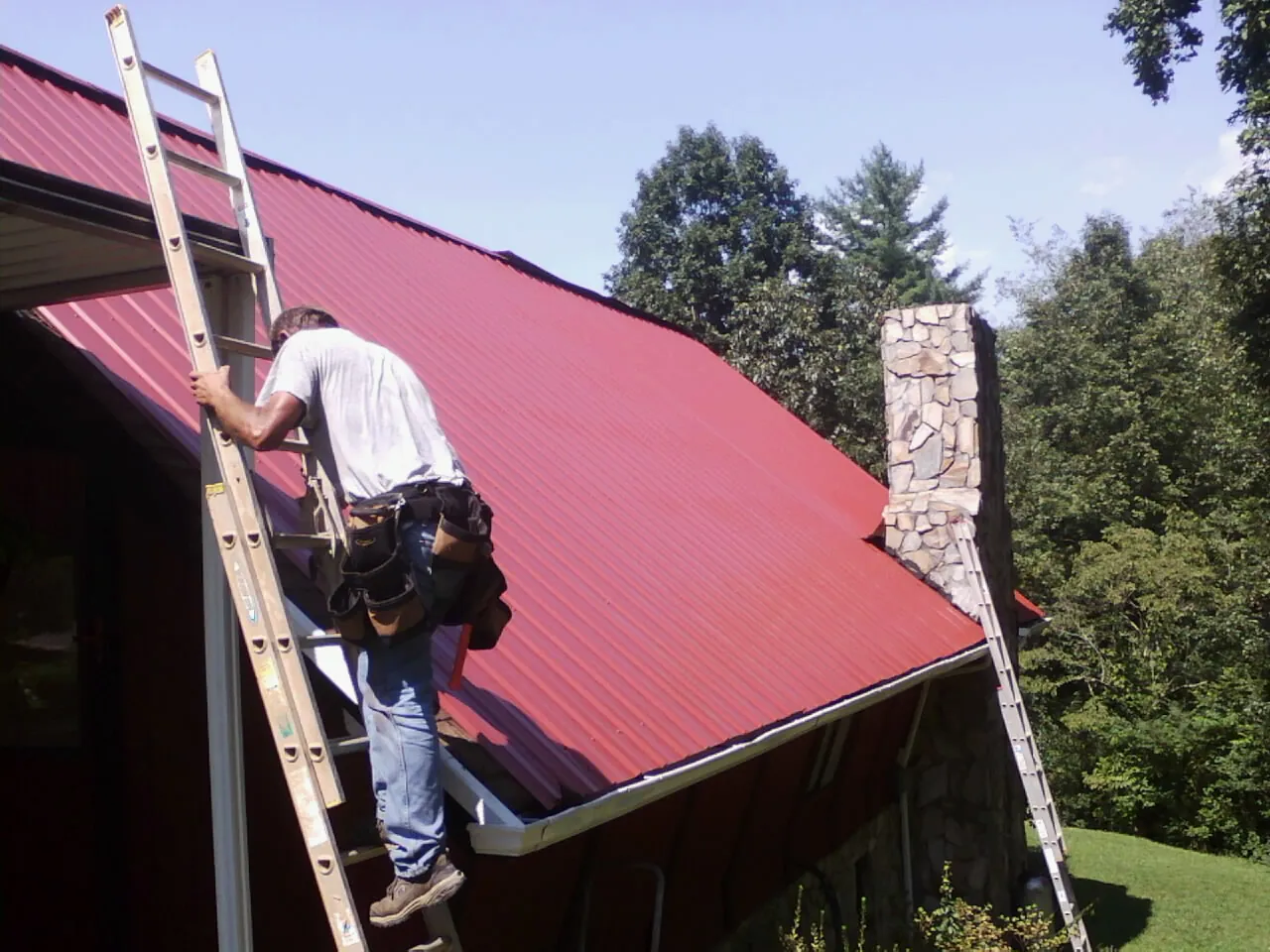 Roofer on a ladder beside a red metal roof and stone chimney — Soffit & Fascia Repair by Roofing Brothers in Duncannon